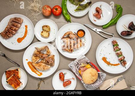 Auf einem Holzkohlegrill auf einem grauen Tisch gegrillte Gerichte. Bratkartoffel, gebratener Chorizo, Hähnchenspieß, Schweinerippchen, Rinderburger. Stockfoto