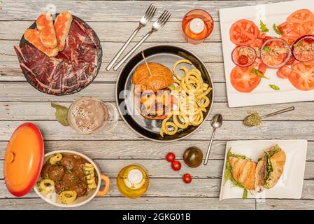 Eine Auswahl an spanischen Gerichten und Hamburgern. Fleischbällchen in Sauce, iberischer Schinken mit Eichel, Tomatensalat, Pastrami-Sandwich Stockfoto