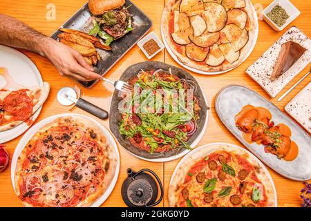 Teller mit spanischen Speisen und italienischen Pizzen von oben betrachtet mit einer Kohlemehlteig-Pizza in der Mitte und einer Männerhand mit einer Gabel bereit, um ca. Stockfoto