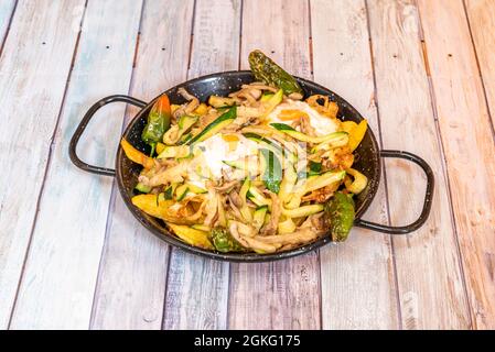 Rühreier mit Gemüse, Zucchini, Paprika, Pilzen und hausgemachten Pommes in schwarz emaillierter Paella-Pfanne auf Holztisch Stockfoto