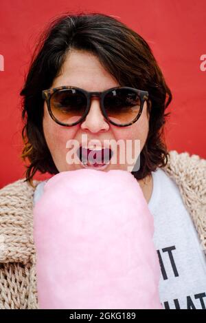 Porträt einer Frau, die gerade dabei ist, einen Bissen aus einer rosa Zuckerwatte zu nehmen. Tragen Sie dunkle Brille und kurze Haare Stockfoto