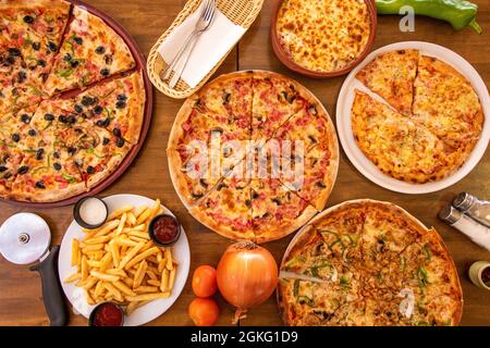 Satz von ganzen Pizzen, die in Portionen mit einigen Zwiebeln, Tomaten und pommes Frites auf dem Holztisch geschnitten wurden Stockfoto