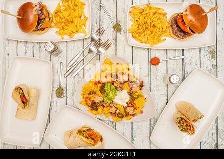 Nachos mit Guacamole, mexikanischen Tacos, Burritos und Hamburgern auf weißem Holztisch mit Gabeln und Löffeln mit Gewürzen Stockfoto