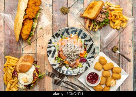 Frisch zubereitetes Essen. Meeresfrüchte-Salat mit Lachs, Aal und Surimi, Thunfisch- und Spargelsandwich, Chicken Nuggets, panierten Hähnchensandwich und Gabeln und Stockfoto