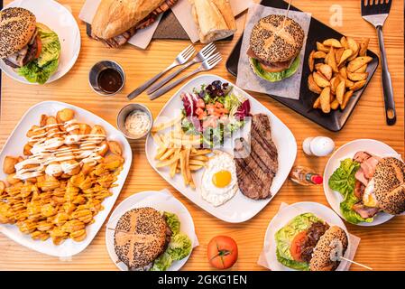 Set mit beliebten spanischen Gerichten und Hamburgern auf Holztisch. Lenden- und Käsesandwich, Steak und Ei, Patatas Bravas, frische Tomaten, Gabeln, deluxe-Zimmer Stockfoto