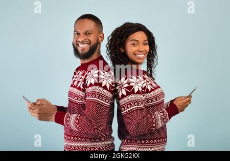 Silvester- und Weihnachtsangebote. Glücklicher afroamerikanischer Mann und Frau, die Smartphones halten und sich zurück an den Rücken lehnen Stockfoto