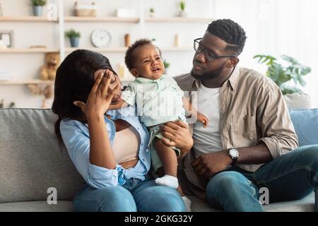 Müde schwarze Eltern sitzen mit weinenden Kind auf dem Sofa Stockfoto