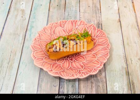 Brioche in Form eines Hot Dogs, gefüllt mit galizisch gekochtem Oktopus auf einem Teller in Form einer rosa Blume Stockfoto