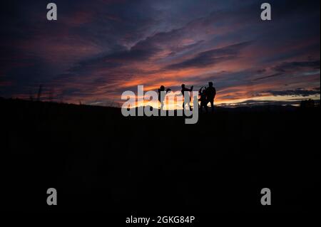 Silhouette einer Familie, die bei Sonnenuntergang mit einem Ball draußen auf einem Hügel unter einem wunderschönen, wolkigen Himmel spielt, der mit den Farben der untergehenden Sonne gefärbt ist. Stockfoto