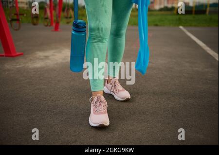 Nahaufnahme. Verkürzte Ansicht der Beine einer sportlichen Frau in Sportkleidung mit einer blauen Plastikflasche Wasser und einem blauen elastischen Gummiband für den Sport Stockfoto