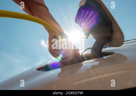 Frau, die das Ladegerät für Elektroautos gegen Sonne und blauen Himmel ansteckt. Stockfoto