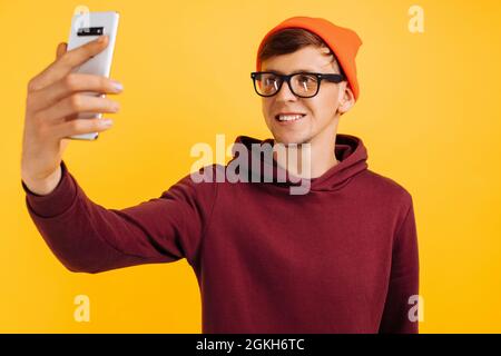 Hübscher junger Mann in einem orangefarbenen Hut mit Brille und einem roten Pullover macht ein Selfie am Telefon, auf gelbem Hintergrund Stockfoto