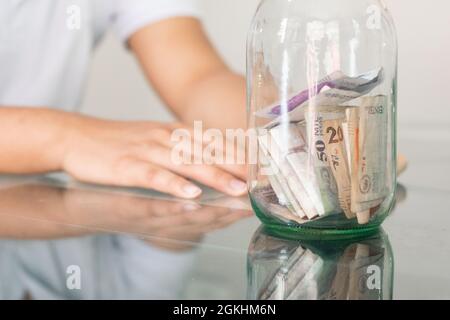 Junge Hand neben seinen Ersparnissen in einem Glas. Junger Mann neben seinen persönlichen Finanzen. Gelangweilter Student mit einer Hand auf dem Kopf. Spar- und Finanzbetrüger Stockfoto