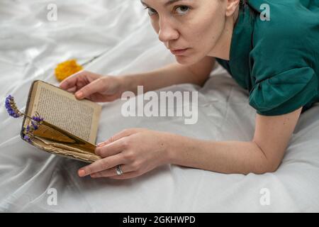Junge Frau mit einem alten Buch und einer getrockneten Lavendelblüte in den Händen, Maniküre mit rosa Nägeln und grünem Hemd auf einem weißen Laken auf einem Kingsize-Bett Stockfoto