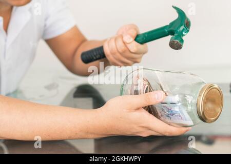 Junger Mann mit Hammer in der Hand bricht seine Sparschwein, Glas voller Banknoten. Spar- und Finanzierungskonzept. Kolumbianische Wirtschaft Stockfoto