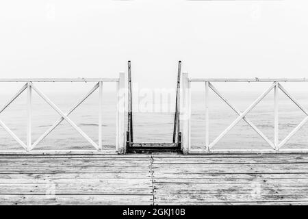 Schwarz-weißes Bild der Promenade mit weißem Geländer mit Treppe, die zum Meer und Holzboden hinuntergeht Stockfoto