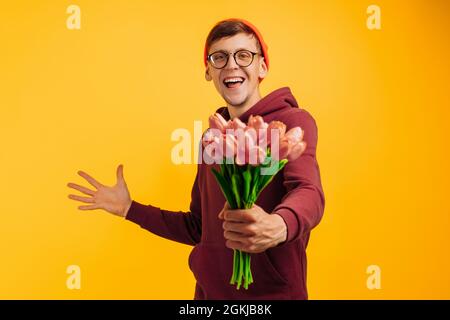 Ein gutaussehender Mann mit einem Blumenstrauß voller Freude gibt Blumen, die in rosa Papier gehüllt sind, ein Mann in einem orangefarbenen Hut und einem roten Pullover mit Blumen in Hi Stockfoto