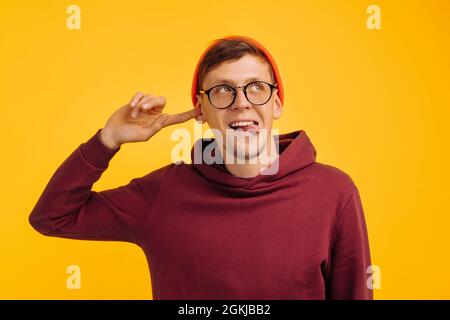 Der Mann bedeckte sein Ohr mit dem Finger, lächelt fröhlich und zeigt seine Zunge, positive Stimmung, in einem orangefarbenen Hut und einem roten Pullover auf gelbem Hintergrund Stockfoto