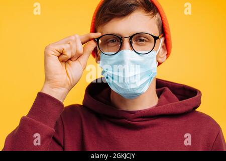 Junger Mann in einer Schutzmaske mit Brille und einem orangen Hut und einem roten Sweatshirt hält seine Hand über die Brille, auf gelbem Hintergrund, Happy vac Stockfoto