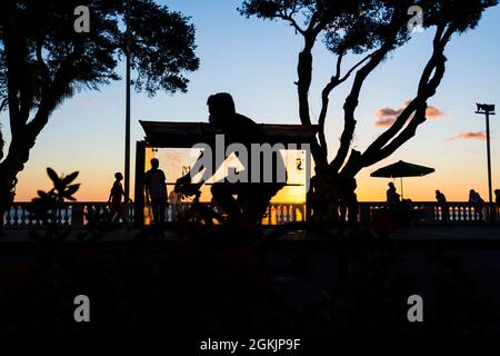 Salvador, Bahia, Brasilien - 17. Juni 2021: Silhouette bei Sonnenuntergang von Baum, Bushaltestelle und Menschen, die am Rande von Porto da Barra spazieren. Stockfoto