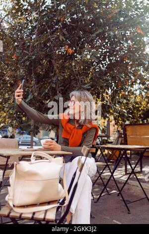 Eine ziemlich ältere Frau macht Selfie mit einem Smartphone, das am Tisch auf der Terrasse des Cafés im Freien sitzt Stockfoto