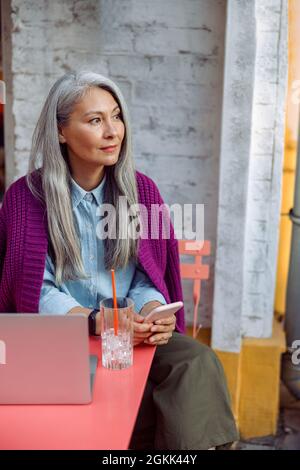 Schöne reife asiatische Frau hält Handy sitzen am Korallentisch mit Notebook Stockfoto
