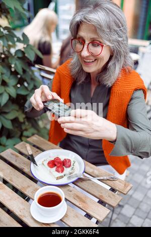 Positive ältere Frau fotografiert köstliche Erdbeerdessert auf der Terrasse des Cafés im Freien Stockfoto