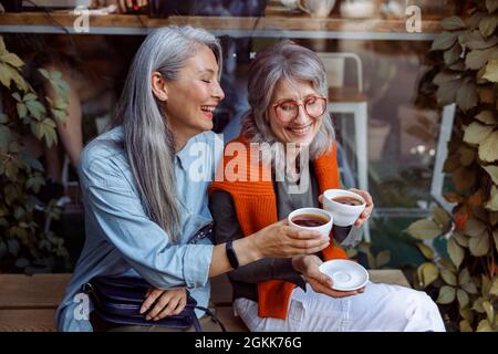 Lächelnde reife asiatische Dame mit Freund clink Tassen Kaffee sitzen in der Nähe des Cafés Stockfoto