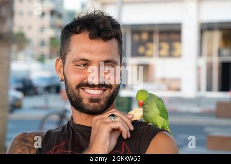Tel Aviv, Israel - 19. August 2021: Ein Mann genießt es, seinen grünen Papagei zu füttern, während er auf seiner Schulter in einer Straße in Tel Aviv steht. Stockfoto