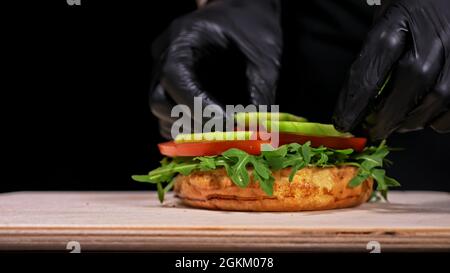 Handwerk Burger ist Kochen auf schwarzem Hintergrund in Schwarz essen Handschuhe. Bestehen: sauce, Rucola, Tomaten, roten Zwiebeln Pommes, Gurken, Käse, Brötchen ein Stockfoto