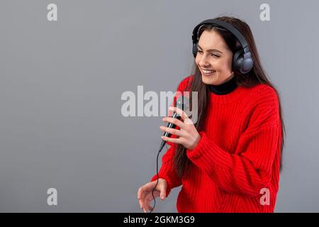 Junge, emotionale Brünette mit Kopfhörern und einem Mikrofon in einem roten Pullover gekleidet singt Karaoke, isoliert auf grau Stockfoto