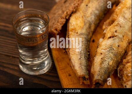 Ein kleiner gebratener Fisch auf einem Holzbrett, Brotscheiben und ein Glas Wodka, Nahaufnahme, selektiver Fokus Stockfoto