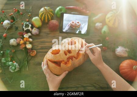 Halloween-Konzept: Weibliche Hände schnitzen Kürbis mit Tutorial auf Tablet. Draufsicht. Frau schnitzt einen Mund in einen Kürbis, um einen Jack o'Laterne auf einem Holztisch mit Gemüse zu machen. Hochwertige Bilder Stockfoto