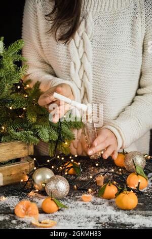 Mädchen in Strickpullover macht ein Weihnachten und legt es in kleine Glasflasche, Mandarinen und Tannenbaum Dekoration hinter Stockfoto