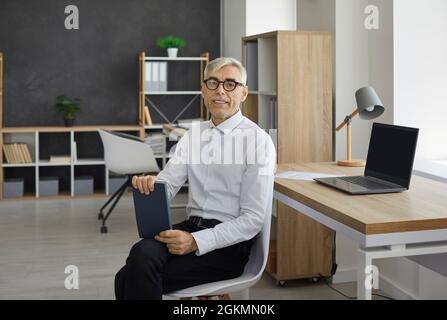 Ein Geschäftsmann, der in seinem Büro auf einem Stuhl sitzt, ein Notebook hält und die Kamera anschaut Stockfoto