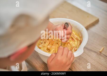 Ein Junge in der Kleidung des Küchenchefs gießt Äpfel in den Teig, um eine Torte zu kochen Stockfoto