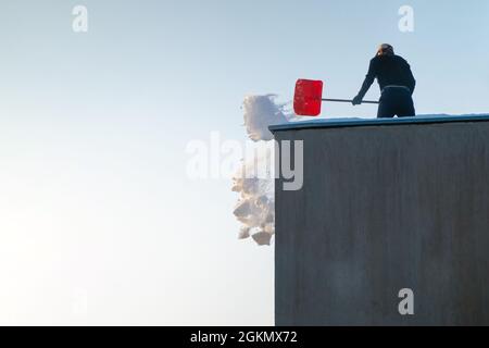 Mann, der an einem sonnigen Wintertag Schnee vom Dach wirft. Arbeiter, die die Oberseite eines Gebäudes reinigen. Stockfoto