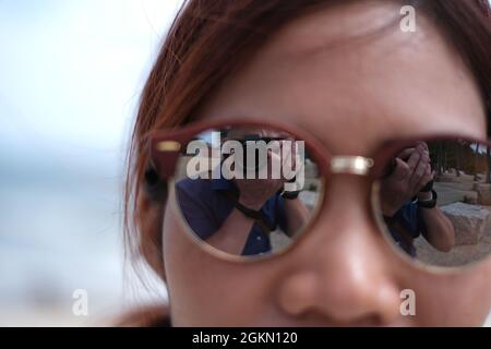 Der Fotograf selbst reflektiert in Sonnenbrillen eine attraktive asiatische Frau Stockfoto