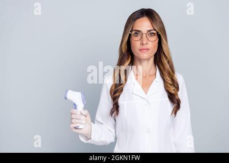 Foto von ziemlich zuversichtlich Dame Arzt tragen weißen Mantel halten berührungslose Thermometer leeren Raum isoliert grauen Hintergrund Stockfoto