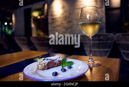 Sizilianische Cannoli Rollen mit Blaubeeren auf einer weißen Keramikplatte und ein Glas Weißwein auf einem Restauranttisch mit Restaurantdekor im Hintergrund Stockfoto