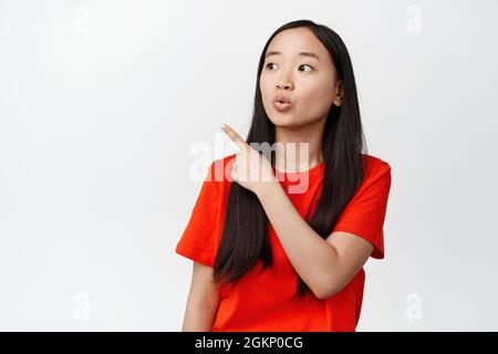 Bild von Brünette asiatische Mädchen Frage stellen, neugierig auf die obere linke Ecke schauen, mit dem Finger auf den Verkauf, Markenlogo, stehend in rotem T-Shirt über Stockfoto