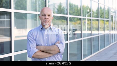 Ein nachdenklicher, selbstbewusster Geschäftsmann, der mit einem intensiven Ausdruck nachdenklich auf die Kamera schaut, während er mit gefalteten Armen vor einem modernen Glas-Co steht Stockfoto