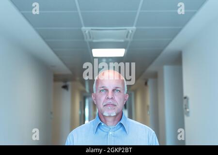 Ein ernsthafter Geschäftsmann mit durchdringenden blauen Augen starrt aufmerksam auf die Kamera, während er durch einen Bürogang in einem Kopf-Schultern-Port geht Stockfoto