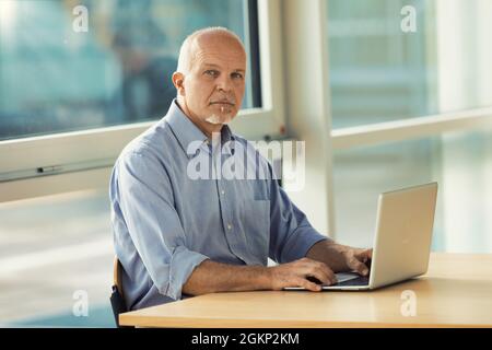 Ein kluger Geschäftsmann, der der Kamera einen einfühlsamen Blick verleiht, während er in einem geräumigen, modernen Büro an einem Laptop arbeitet Stockfoto