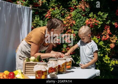 Konservierung Gartengemüse, Konservierung von Tomaten, Paprika, Zucchini-Gemüse. Mama und Sohn Gemüsekonserven in Gläsern auf dem Tisch in der Stockfoto