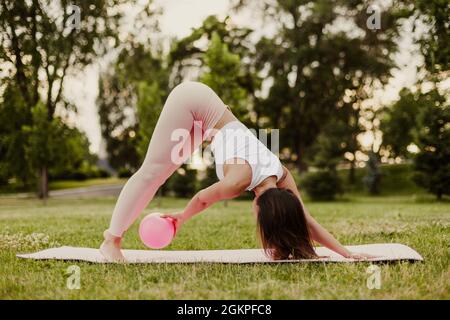 Eine athletische, schlanke Frau in rosa Kleidern und ein kleiner Gummiball, die im Sommer im Park Pilates auf einer rosa Matte bei Sonnenaufgang macht Stockfoto