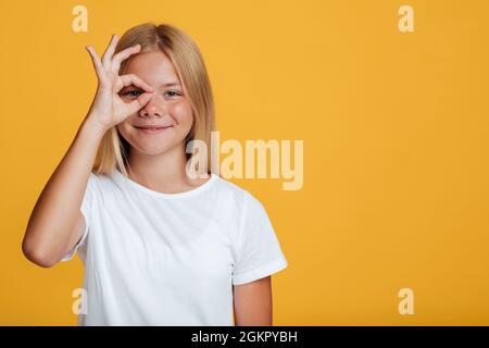 Glücklich hübschen Teenager blonde Dame Schüler in weißem T-Shirt zeigt ok Geste nahe Augen Stockfoto