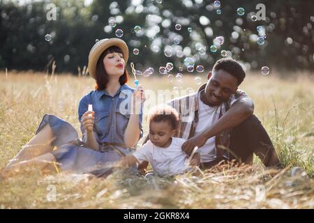 Schöne kaukasische Mutter und hübscher afrikanischer Vater blasen Seifenblasen während des Sommerpicknicks mit kleinen Sohn auf die Natur. Familie, die Spaß im Freien hat. Entspannungskonzept. Stockfoto