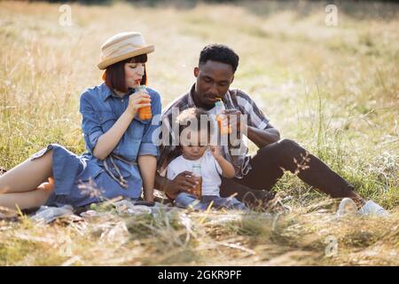 Positive junge Eltern und ihr süßer Sohn sitzen zusammen auf Gras und trinken frischen Saft aus Stroh. Entzückende multiethnische Familie genießt Picknick-Zeit während der Sommertage. Stockfoto