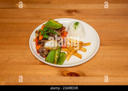 Peruanischer Lomo Saltado mit Gemüse, hausgemachten Chips und Schüssel mit weißem Reis mit Olivenöl und Petersilie auf weißem Teller Stockfoto